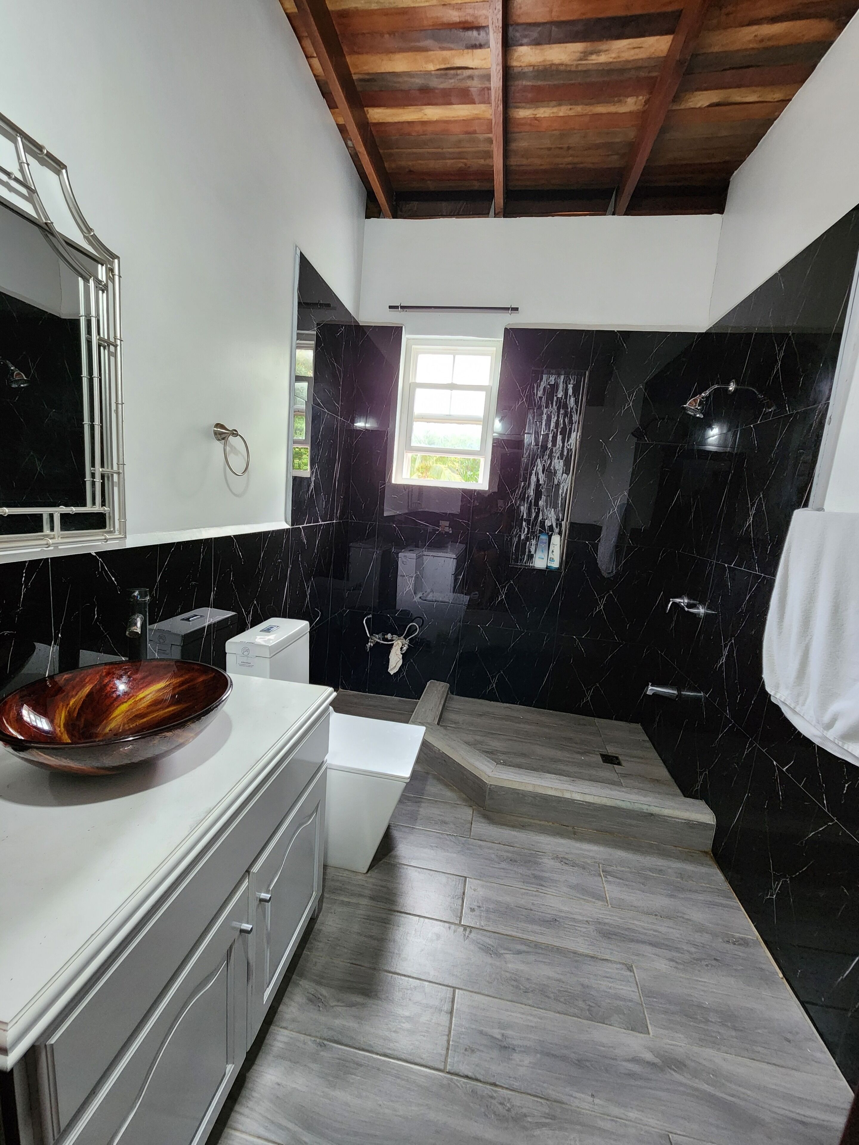 Un baño moderno con techo de madera, un espejo grande, un lavamanos de mármol negro y encimera blanca.