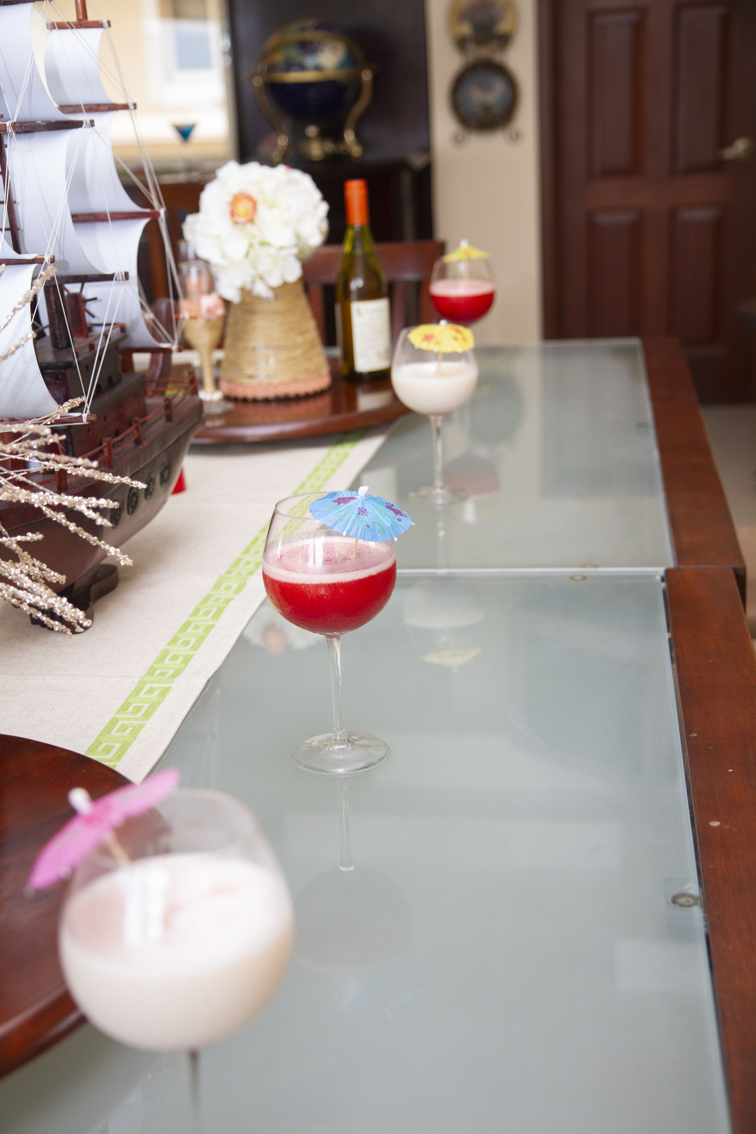 A dining table with three glasses of red drinks, a ship model, and a vase of flowers.