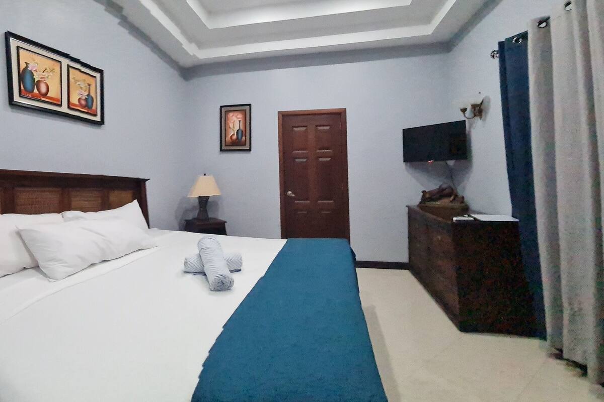 Una cama bien hecha con sábanas blancas y un detalle azul, un cabecero de madera, una mesita de noche con una lámpara, un televisor sobre un mueble, y una puerta con cortinas.
