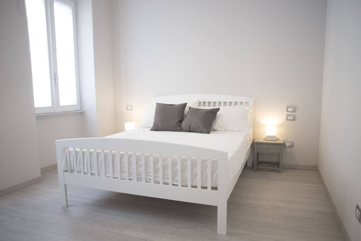 A white bed with a wooden headboard and two pillows, a nightstand with a lamp, and a window with blinds.