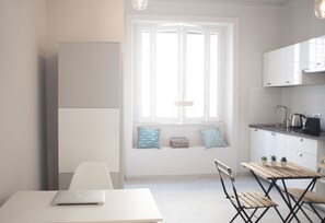 Una cocina moderna con armarios blancos, un ventanal con persianas, una mesa de comedor con sillas y un ordenador portátil sobre la mesa.