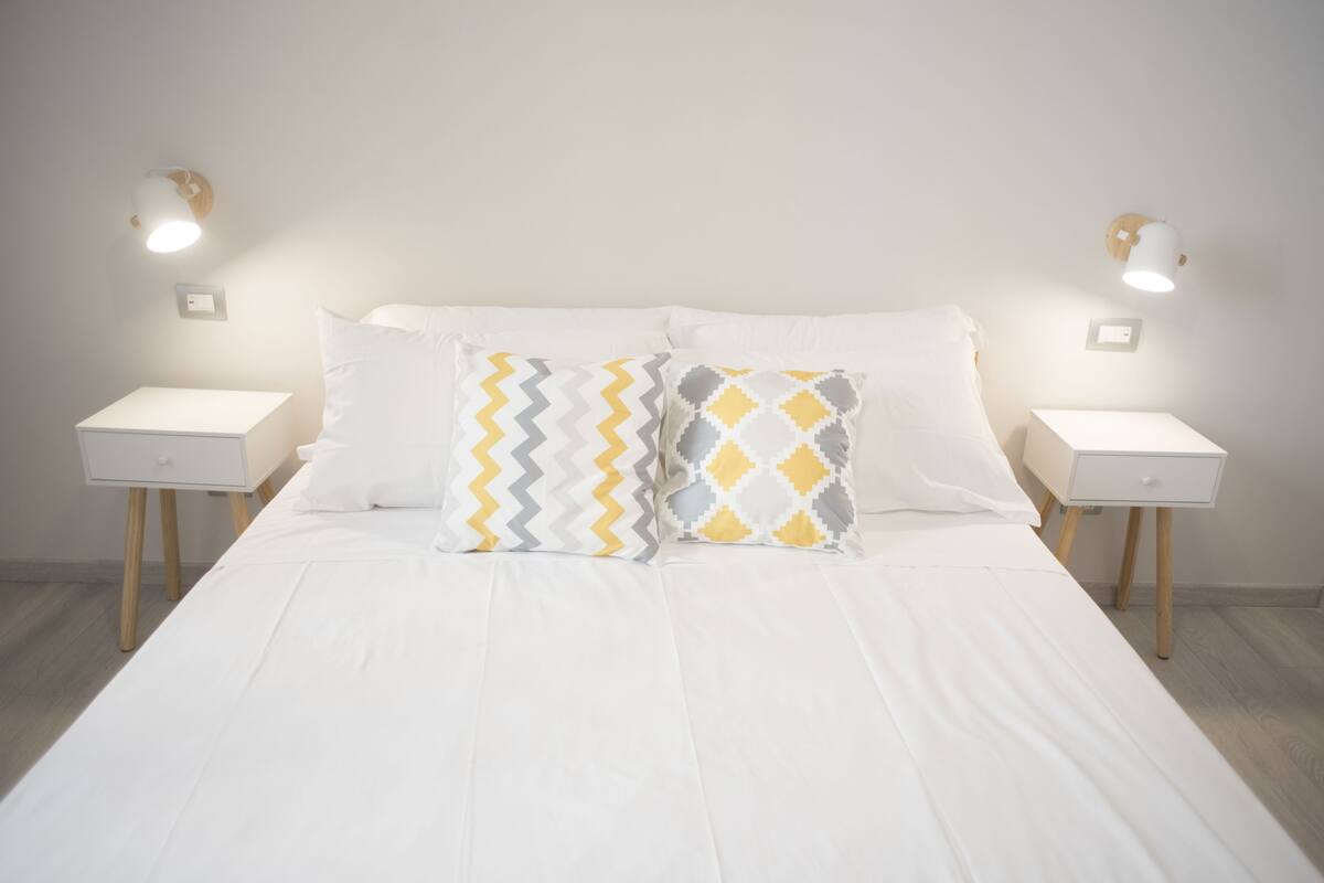 A neatly made bed with white bedding and two patterned pillows, flanked by bedside tables with lamps, in a minimalist room with light wooden flooring.