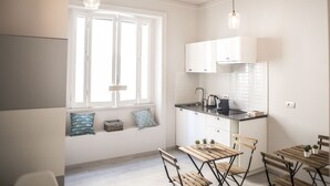 Una cocina moderna con armarios blancos, encimera negra y sillas de madera. Hay una ventana con persianas, una lámpara colgante y una pequeña planta en la encimera.