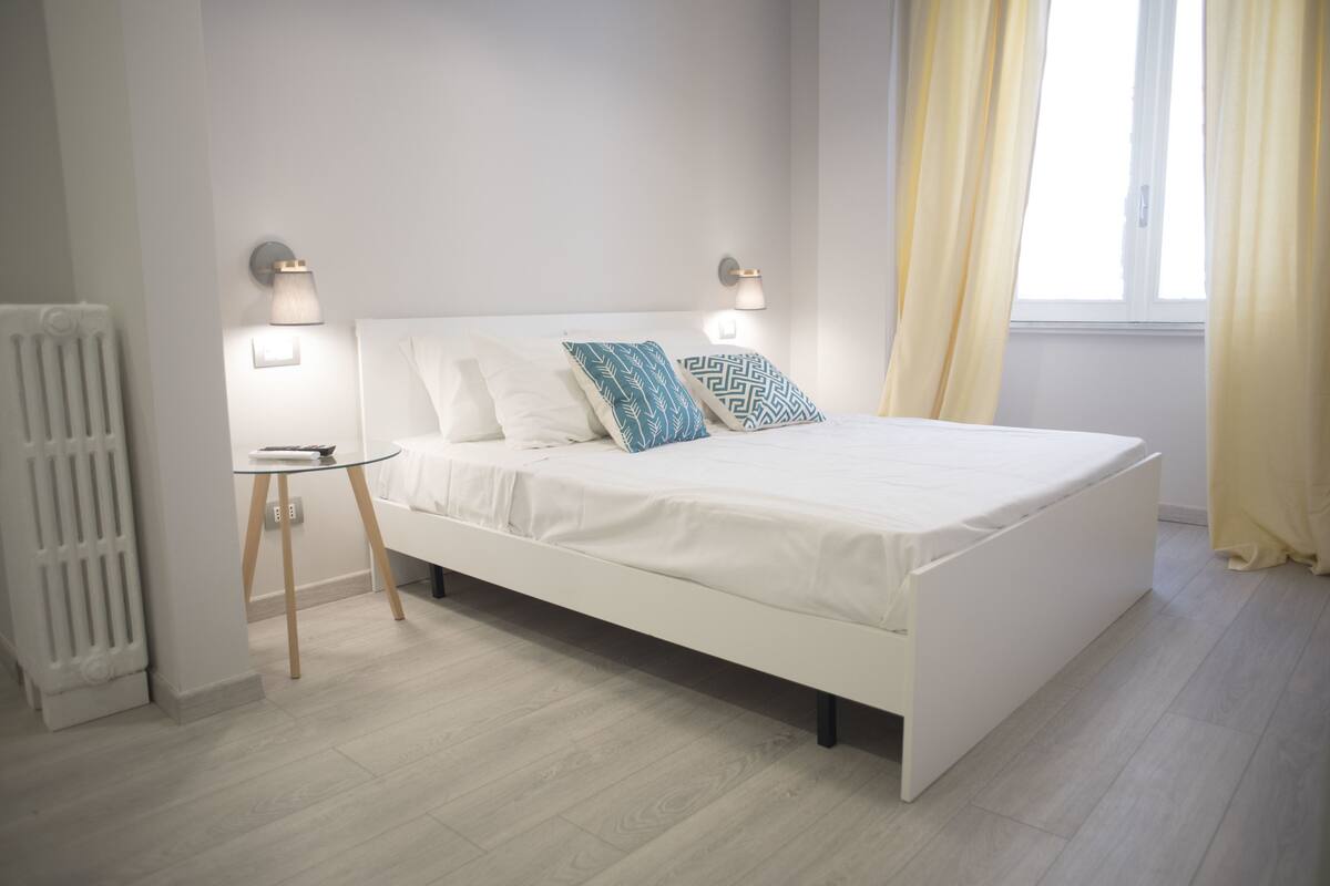 A modern bedroom with a white bed, two decorative pillows, a small bedside table, and a wall-mounted lamp.
