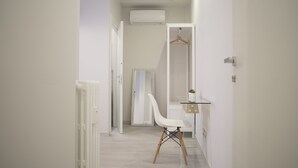 Un pasillo estrecho con un radiador blanco, una silla blanca y una mesa de vidrio con una planta colgante.