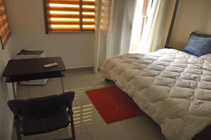 A hotel room with a bed, a desk, a chair, and a red welcome mat.