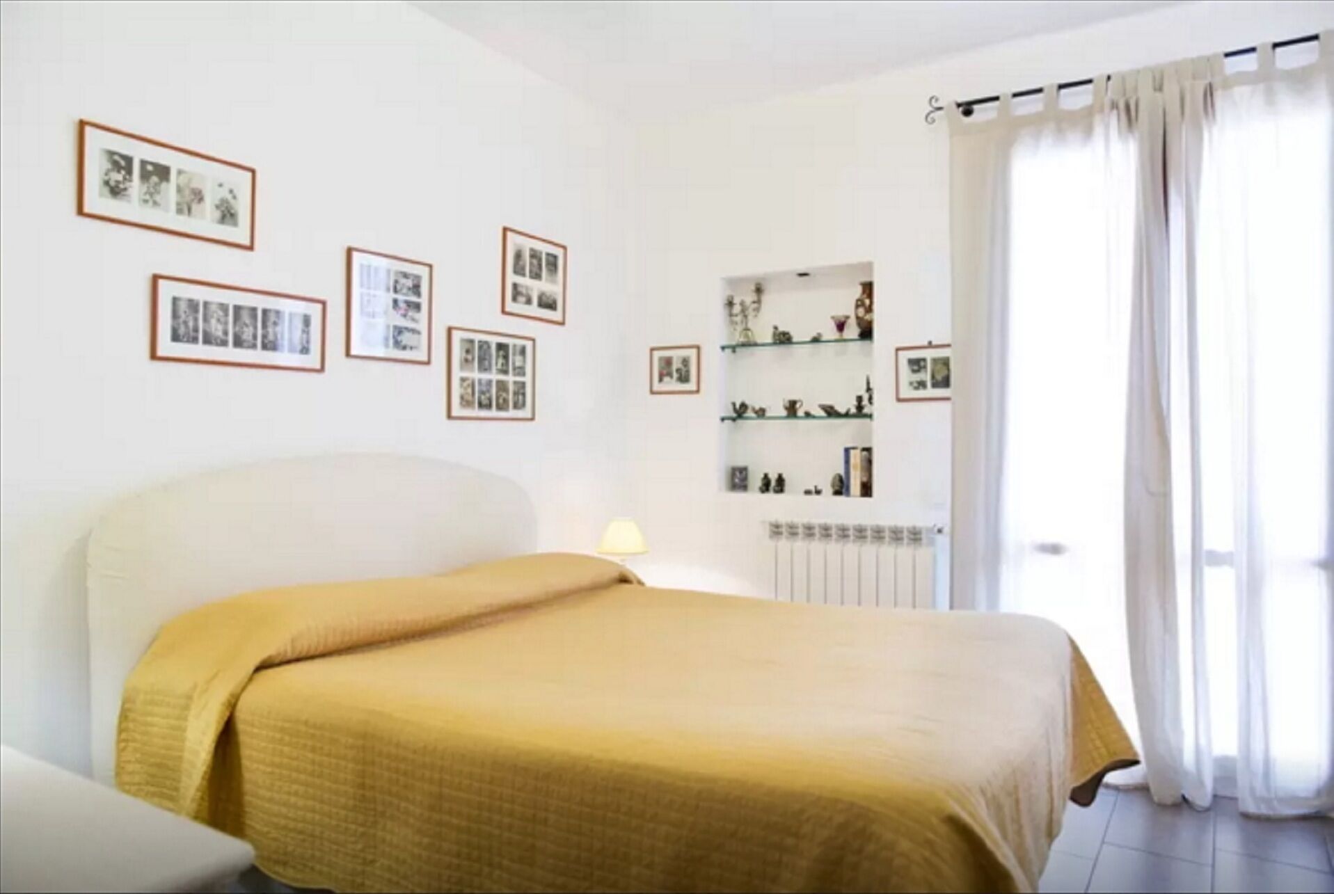 A bedroom with a bed, a bedside table, a lamp, a radiator, and a window with curtains.