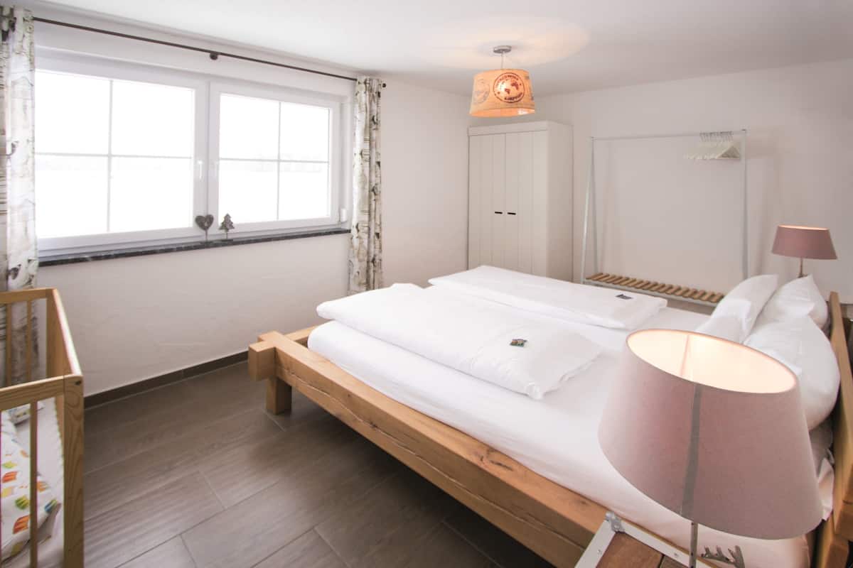 A bedroom with a wooden bed, white bedding, a bedside lamp, a wardrobe, and a window with curtains.