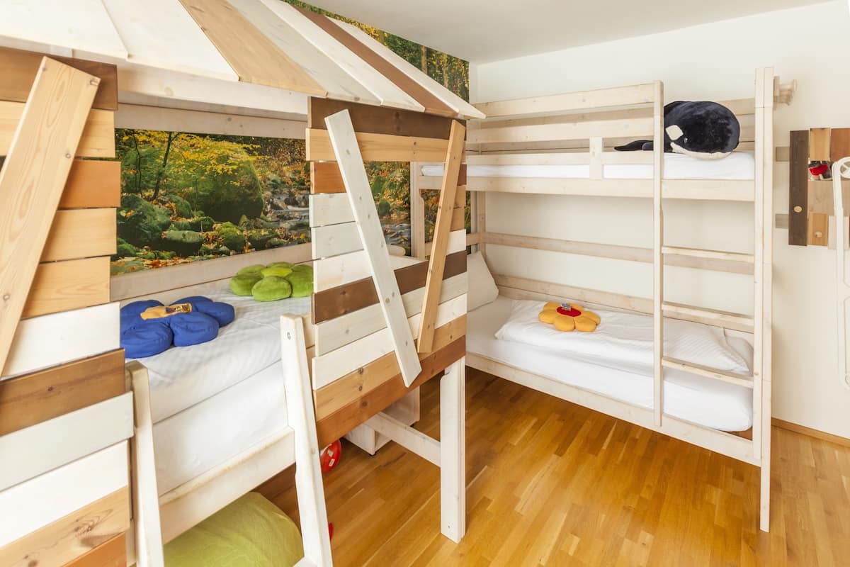 A bunk bed room with a wooden ladder, a green mat, and a small window with a view of trees.