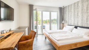 Ein Hotelzimmer mit einem hölzernen Bett, einem Schreibtisch mit Stuhl, einem an der Wand befestigten Fernseher und einem Balkon mit Blick auf Bäume und Gebäude.