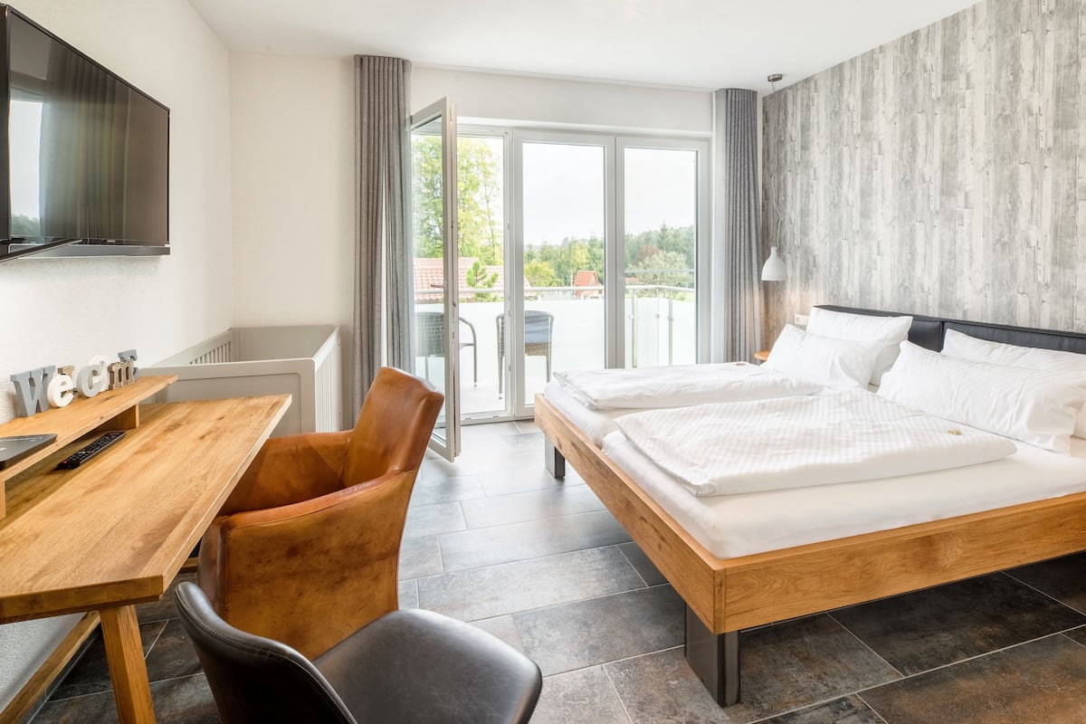 A hotel room with a wooden bed, a desk with a chair, a TV mounted on the wall, and a balcony with a view of trees and buildings.