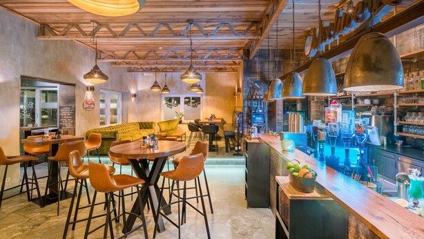 Eine gemütliche Bar mit Holzbalken, hängenden Pendelleuchten und einer Theke mit verschiedenen Flaschen und Gläsern.