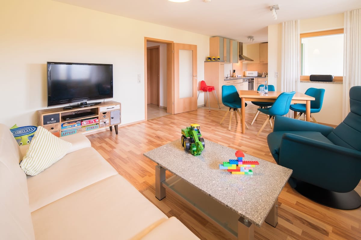 A modern living room with a flat-screen TV, a sofa, a coffee table with toys, and a dining area with blue chairs.