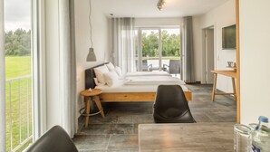 Ein modernes Schlafzimmer mit einem großen Bett, einem Schreibtisch und einem Stuhl. Durch das Fenster ist ein Blick auf eine grüne Landschaft möglich.