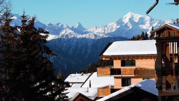 A wooden chalet with a balcony, set against a backdrop of snow-covered mountains and clear blue sky.