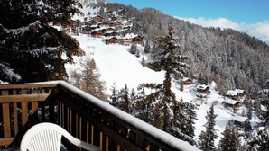 A snow-covered mountain village with wooden chalets, a wooden deck with a white plastic chair, and a clear sky.