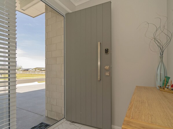 Un hall d’entrée moderne avec une porte grise, une petite fenêtre avec des stores et une table en bois sur laquelle est posé un vase.