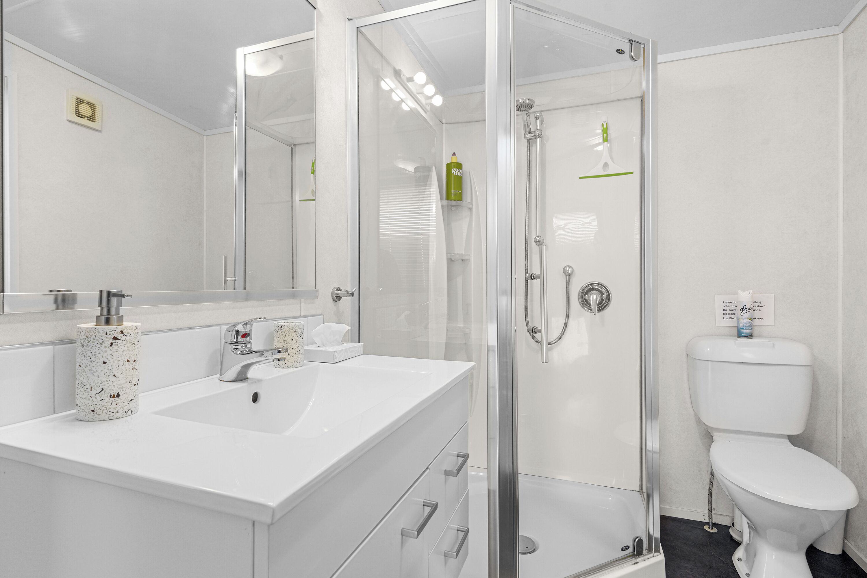 A modern bathroom with a glass-enclosed shower, a white sink with a speckled soap dispenser, and a toilet with a green bottle on the wall.