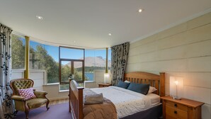 A bedroom with a large bed, a wooden nightstand, a chair, and a view of mountains through a large window. - Lakes Edge Apartment - Unit A (Queenstown)