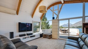 A modern living room with a large flat-screen TV, leather sofas, and a view of mountains through large windows. - Lakes Edge Apartment - Unit A (Queenstown)