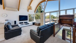 A modern living room with a flat-screen TV, leather sofas, and a large window offering a view of the outdoors. - Lakes Edge Apartment - Unit A (Queenstown)