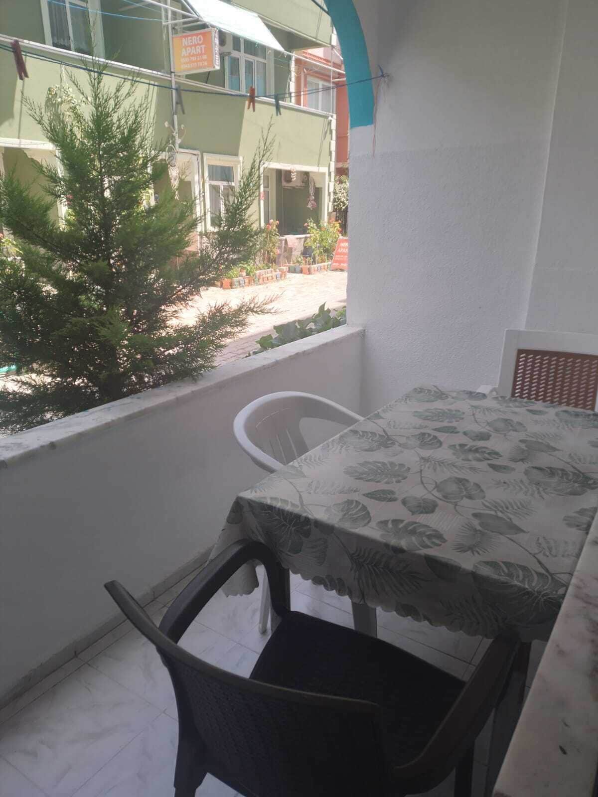 A balcony with a table and chairs, a small tree, and a building with a sign in the background.