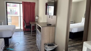 Ein Hotelzimmer mit einem Bett, einem Schreibtisch mit Stuhl, einem Fernseher, einem Mülleimer und einer Schiebetür zum Balkon.