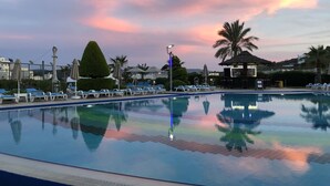 Ein Schwimmbecken mit Liegestühlen und Sonnenschirmen, umgeben von Palmen und einem Abendhimmel.