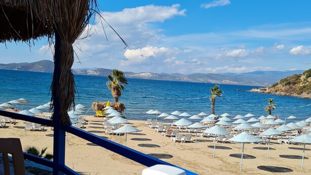 Blick auf einen Strand vom Balkon aus, mit einem Holztisch, weißen Hockern und Sonnenschirmen.