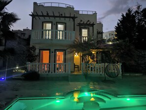 Een gebouw met twee verdiepingen, een balkon, een zwembad met groene verlichting en een goed verlichte ingang.