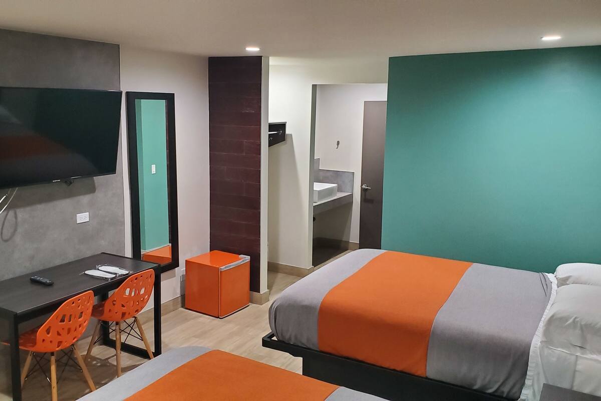 A modern hotel room with a bed, a desk, two orange chairs, a television, and a bathroom visible through an open door.