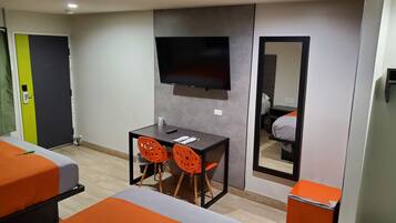 Habitación de hotel con una cama, un escritorio con dos sillas, un televisor de pantalla plana en la pared y un espejo que refleja la habitación.