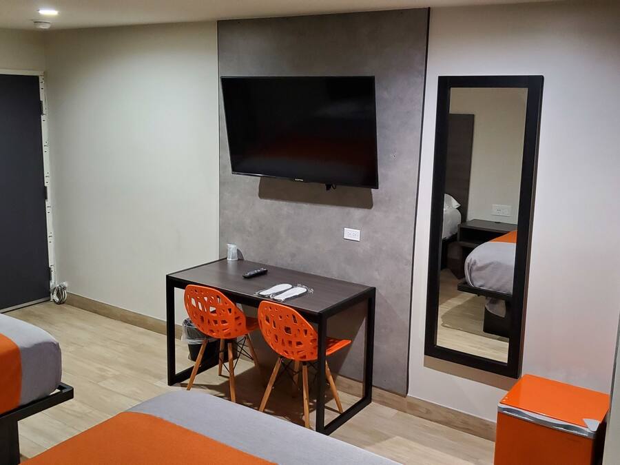 Habitación de hotel con una cama, un escritorio con dos sillas, un televisor de pantalla plana en la pared y un espejo que refleja la habitación.