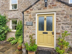 Une maison en pierre avec une porte jaune, une enseigne indiquant « Corner House », et des plantes en pot disposées autour de l’entrée.
