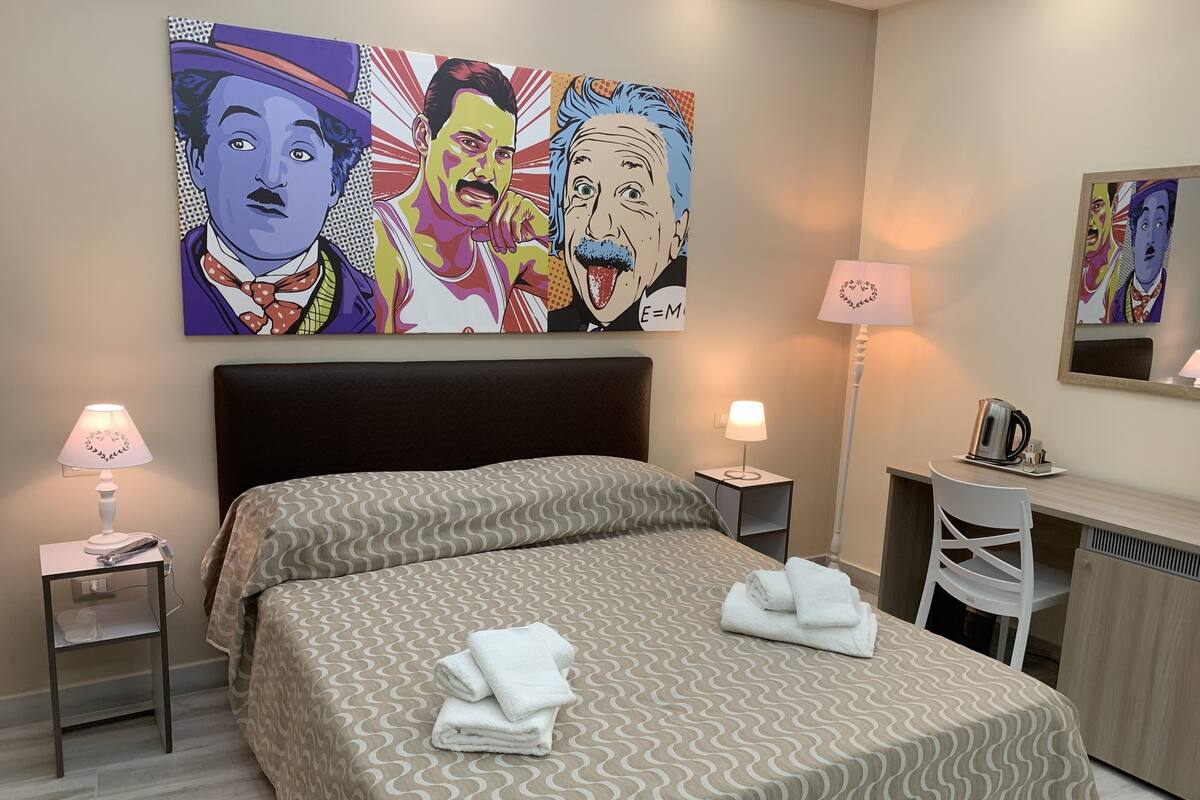 A hotel room with a bed, two bedside tables, a desk, and a chair. There are lamps on the bedside tables and a kettle on the desk.