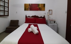 Una cama bien hecha con ropa de cama roja y blanca, un cabecero de madera y una manta roja. Hay una mesita de noche pequeña con una lámpara y un cuadro enmarcado en la pared.