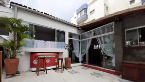 Una furgoneta camper roja marca VW estacionada frente a un edificio con ventanas amplias y un acceso de vidrio. Hay dos taburetes de madera y una planta en maceta cerca de la furgoneta.