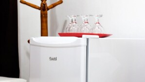 Un refrigerador blanco con una bandeja roja encima, una silla de madera y una cama blanca al frente.