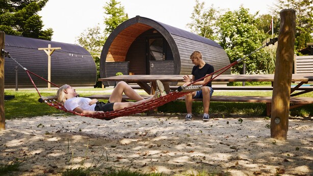 Two people relaxing on a hammock between two cabins with a wooden bench in the foreground.