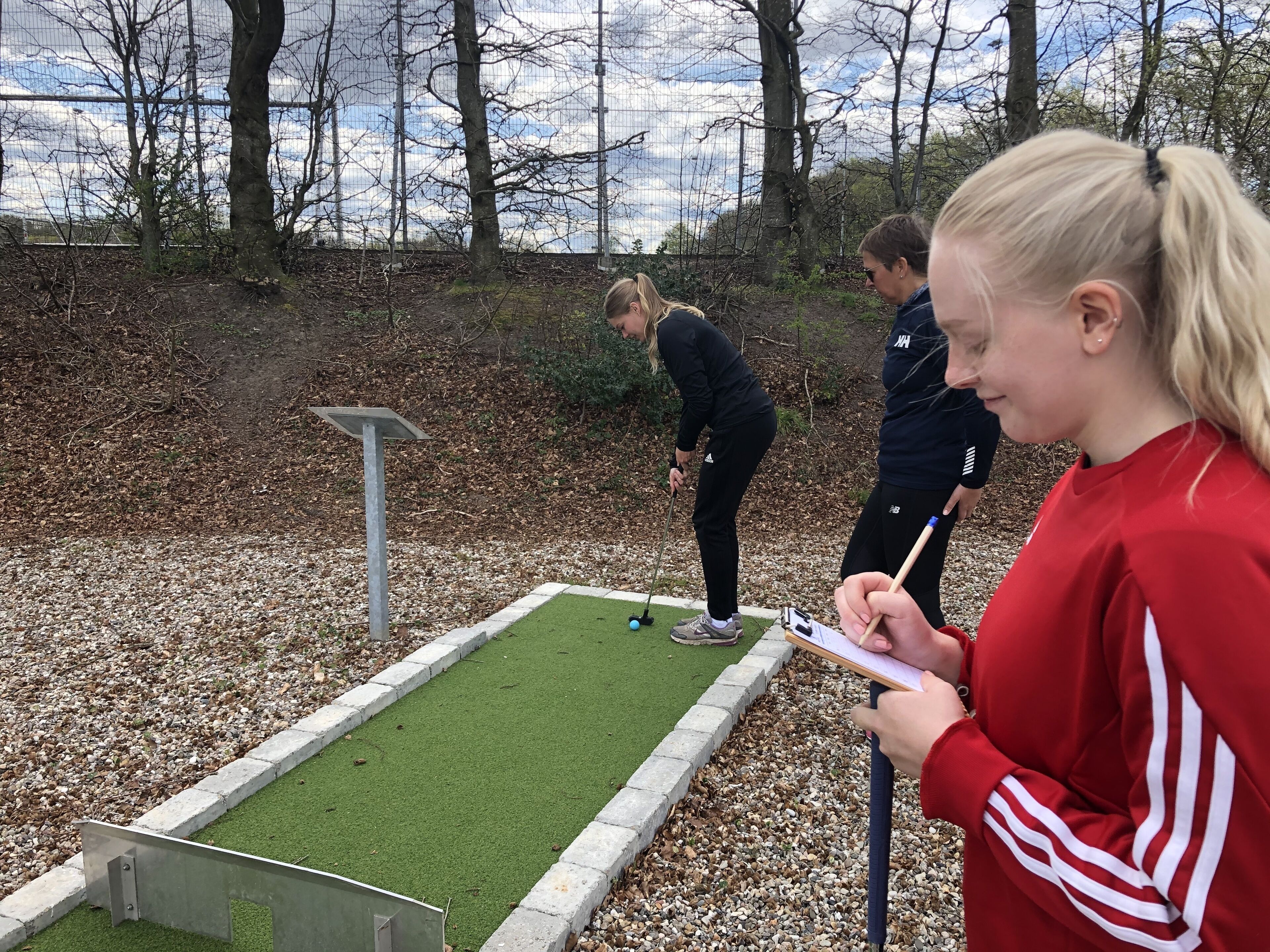 Una persona con una chaqueta roja tomando notas cerca de un campo de mini golf, con un putting green y una barrera de metal.