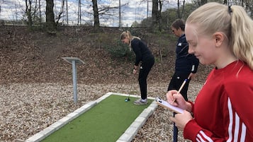 Una persona con una chaqueta roja tomando notas cerca de un campo de mini golf, con un putting green y una barrera de metal.
