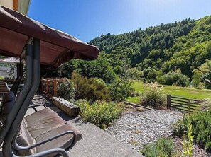 Eine Terrasse mit Sitzgelegenheit, Blick auf einen bewaldeten Hügel und ein Kiesweg, der zu einem Holzzaun führt.