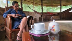 A person sitting on a wooden chair in a room with a thatched roof, a hammock, and a table with a cup and a decorative pot. - Intillama Jungle Lodge (Fernando Lores)