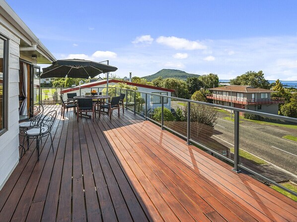 A wooden deck with outdoor furniture, a glass balustrade, and a view of a road and hills.