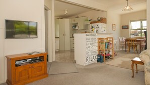 A living room with a wooden cabinet, a bookshelf, and a dining area with chairs and a table. - Surf Beach Views - Whangamata Holiday Home (Whangamata)