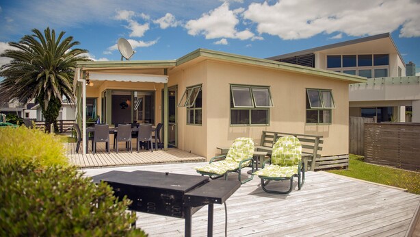A house with a patio area featuring a table and chairs, a satellite dish, and a palm tree. - Surf Beach Views - Whangamata Holiday Home (Whangamata)