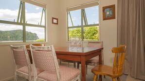 A dining area with a wooden table and chairs, large windows with a view of greenery and hills, and a framed picture on the wall. - Surf Beach Views - Whangamata Holiday Home (Whangamata)