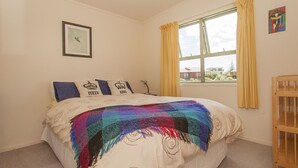 A bedroom with a bed, a window with curtains, a picture on the wall, and a small wooden cabinet. - Surf Beach Views - Whangamata Holiday Home (Whangamata)