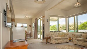 A spacious living room with large windows, a wooden desk, and a sofa. - Surf Beach Views - Whangamata Holiday Home (Whangamata)