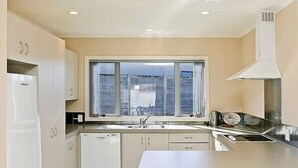 Cocina compacta con gabinetes blancos, horno integrado, lavamanos y ventana con persianas.